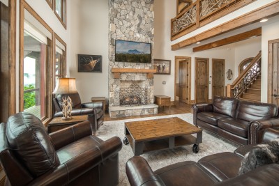The great room rises two stories to exposed timber beams, anchored by a dry-stacked stone chimney, a lit gas fire, and enough leather seating for all twelve of you.