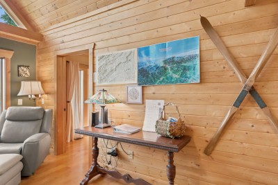 Pine plank wall with a raised relief map and a painted aerial view of the mountains, crossed vintage wooden skis, a carved wood console table with stained glass lamp, binder and woven basket, and a gray recliner in a doorway.