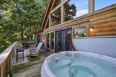 The hot tub sits just outside the great room's glass doors, so it is a few barefoot steps from the sofa to the water. Rocking chairs and an outdoor bar table share the deck, along the log wall and soaring windows on the view side of the cabin.