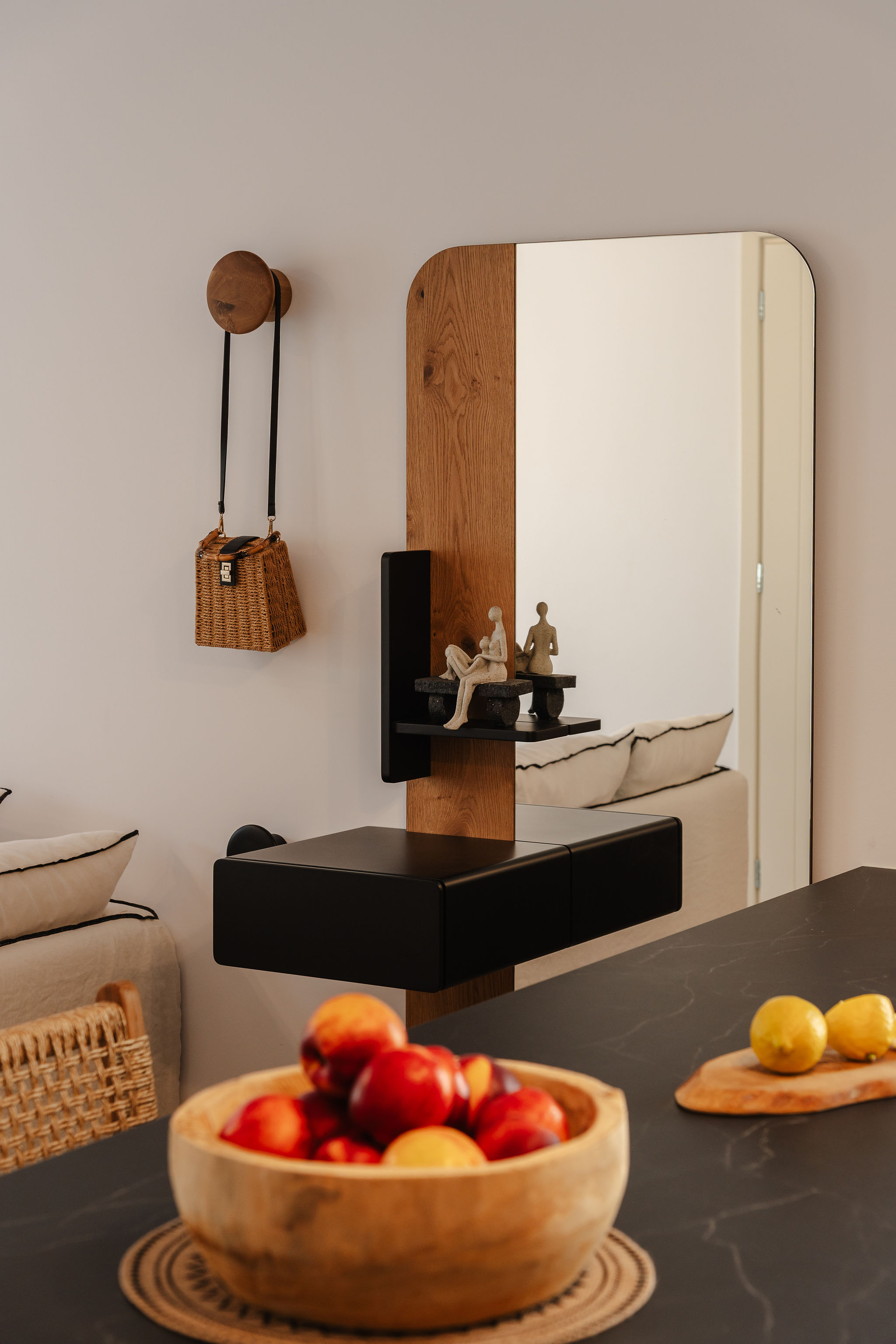 A stylish entryway or dining area featuring a tall mirror mounted on a wood panel, a floating black console, woven decor, and a bowl of fruit on a dark stone table.