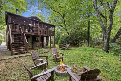 The backyard opens into a wide grassy clearing where Adirondack chairs ring the fire pit and a picnic table sits nearby — beyond the grass, a quiet creek runs along the tree line under a canopy of tall hardwoods and the screened deck above.