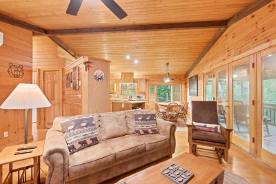 From the sofa, the whole cabin unfolds in one line of sight — the round dining table, the kitchen beyond it, and glass French doors opening straight onto the screened porch and its row of rockers.