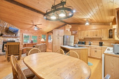 Vaulted pine ceilings stretch over the whole great room, tying the oval dining table under its iron ring chandelier to the oak kitchen island and the stone fireplace beyond — one open space where the whole group ends up together.