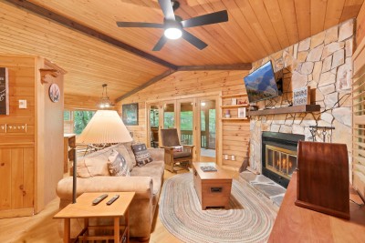 This side of the living room opens through French doors onto the screened porch and its tree-lined view, with the stone fireplace, mounted TV, and mission rocker all within reach of the deep, pillow-piled sofa.