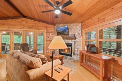 Wood-blind windows frame the trees on one side and open French doors lead to the porch on the other, with a corner stone fireplace, a vintage radio on the console, and plenty of room to stretch out on the sofa.