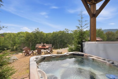 Steam rises off the hot tub while the fire pit's Adirondack chairs and a neighboring cabin roof sit just below, a forested valley stretching out beyond.