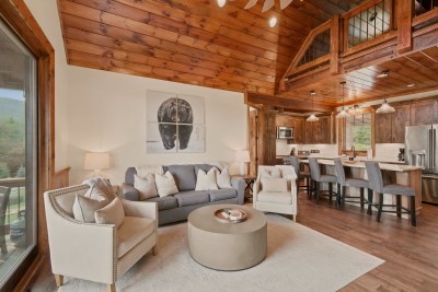 The living area flows straight into the kitchen beneath the vaulted, timber-loft ceiling, gray sofa and nailhead chairs facing the island's barstool seating.