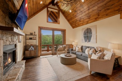 The vaulted great room opens straight onto the deck through glass doors, stone fireplace and wall-mounted TV facing the sofa seating, forested ridgeline visible beyond the rail.
