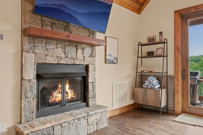 A reclaimed-wood mantel tops the stacked-stone fireplace, with the glass door right beside it opening straight onto the deck and gas grill for easy outdoor access.