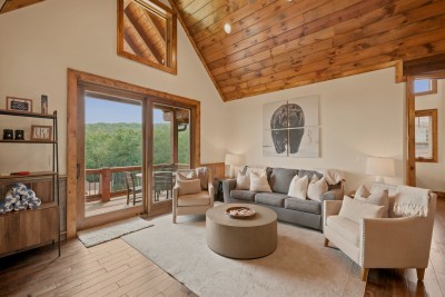 Glass doors slide open onto a deck with its own dining table, wooded hillside rolling out beyond the rail, right past the gray sofa and nailhead chair seating.