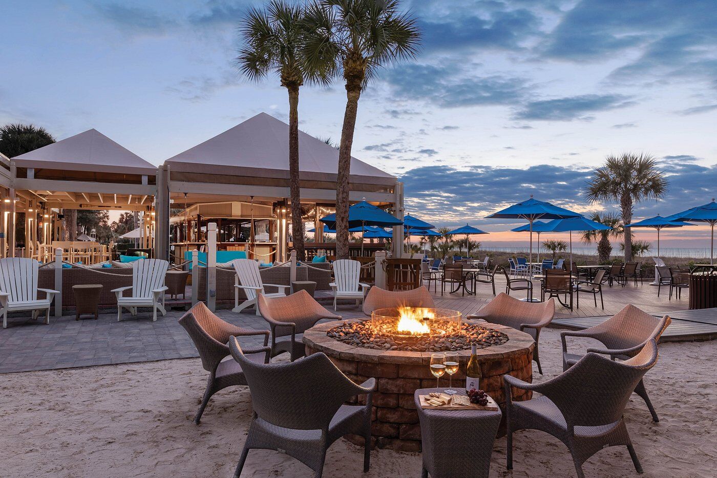 Sunset waterfront lounge with fire pit, palm trees, and resort-style seating