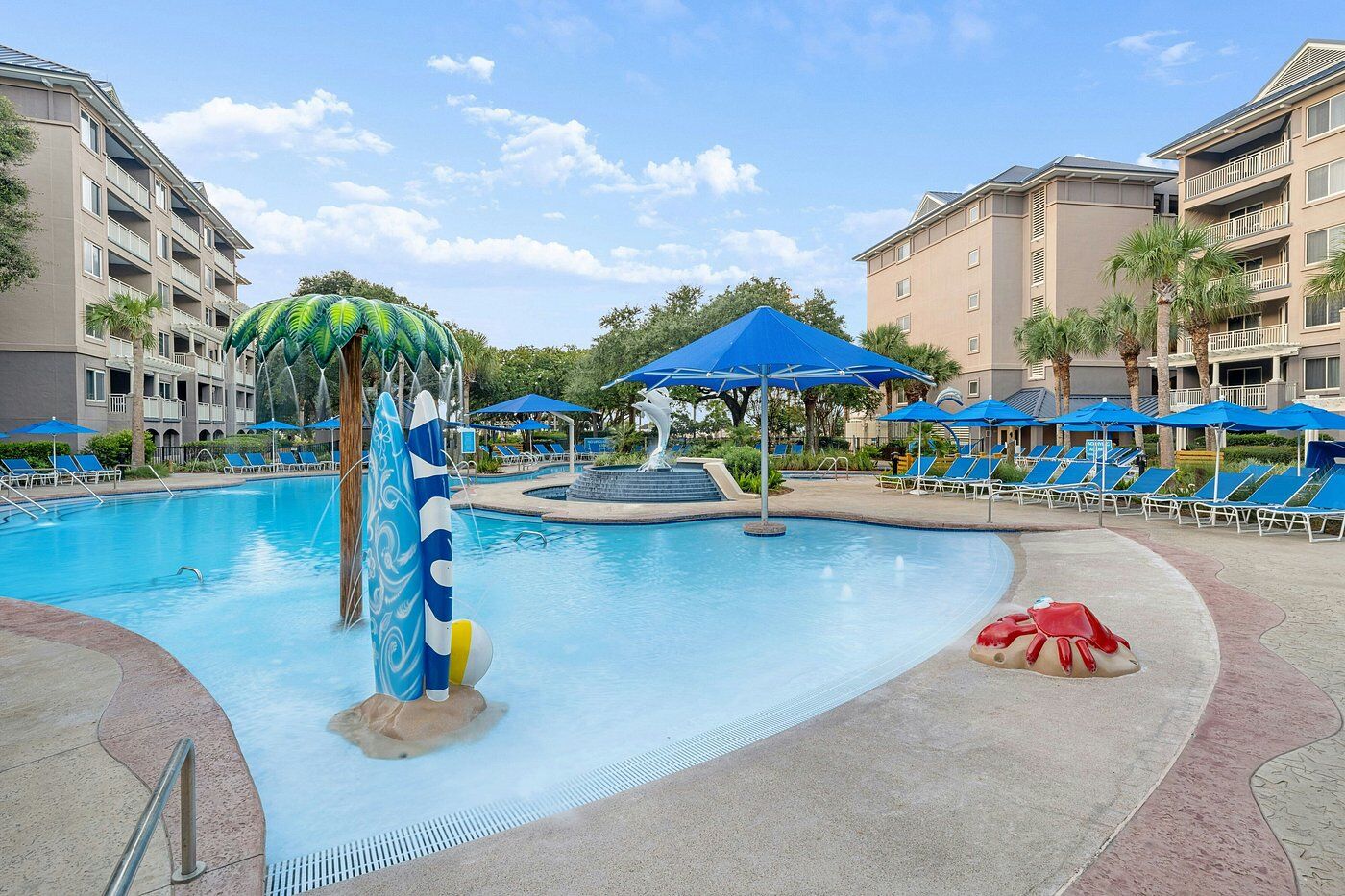 Resort-style pool with splash features, lounge chairs, and sunny palm-lined views