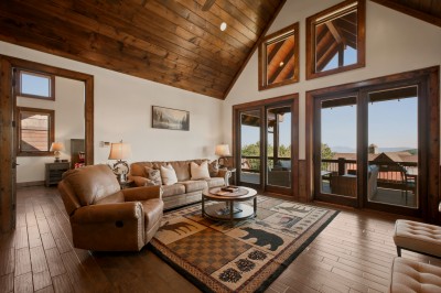 Curl up on plush leather seating beneath soaring wood-plank ceilings, with wall-to-wall windows opening onto endless mountain vistas — a wildlife-patterned rug and warm textures complete this alpine living space.