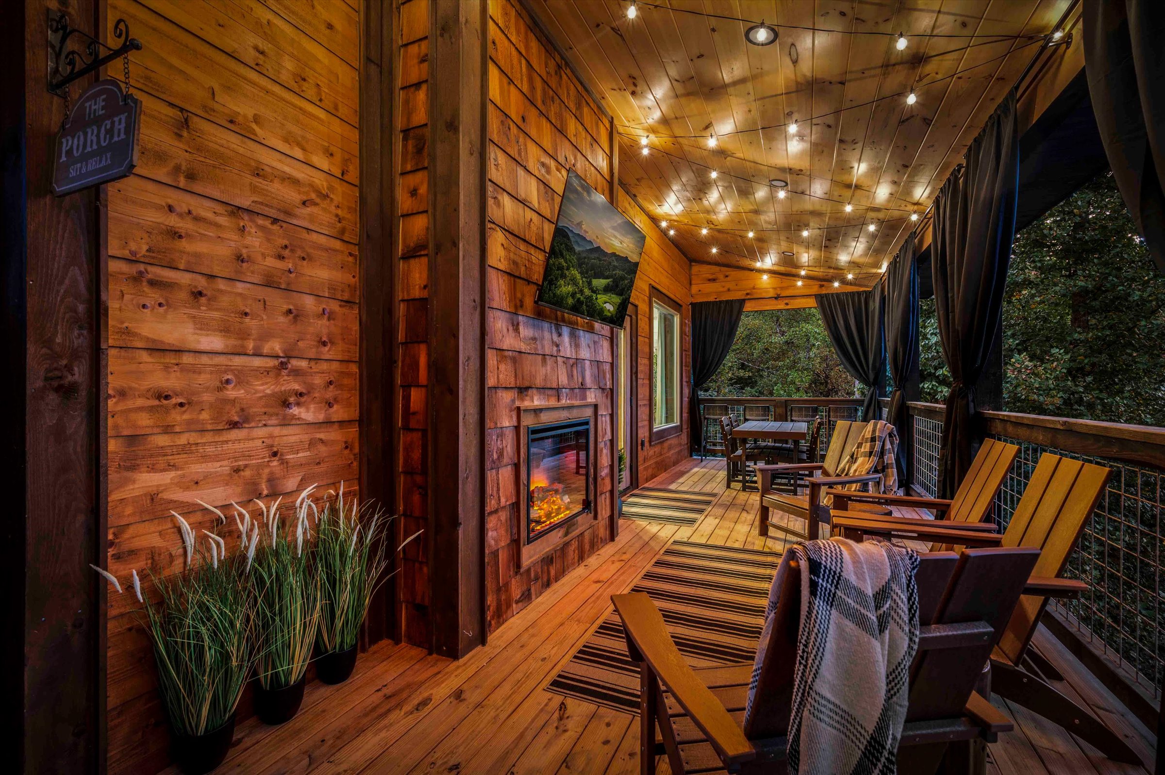 Cozy covered porch with fireplace, TV, string lights, and forest views