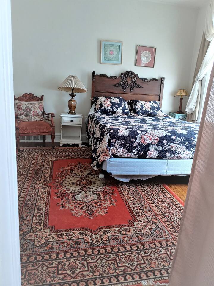 Same bedroom, but with a queen size bed now! The bedroom is spacious with a view of the street, a seating area, and a desk with a chair, and flat screen Tv in the room.