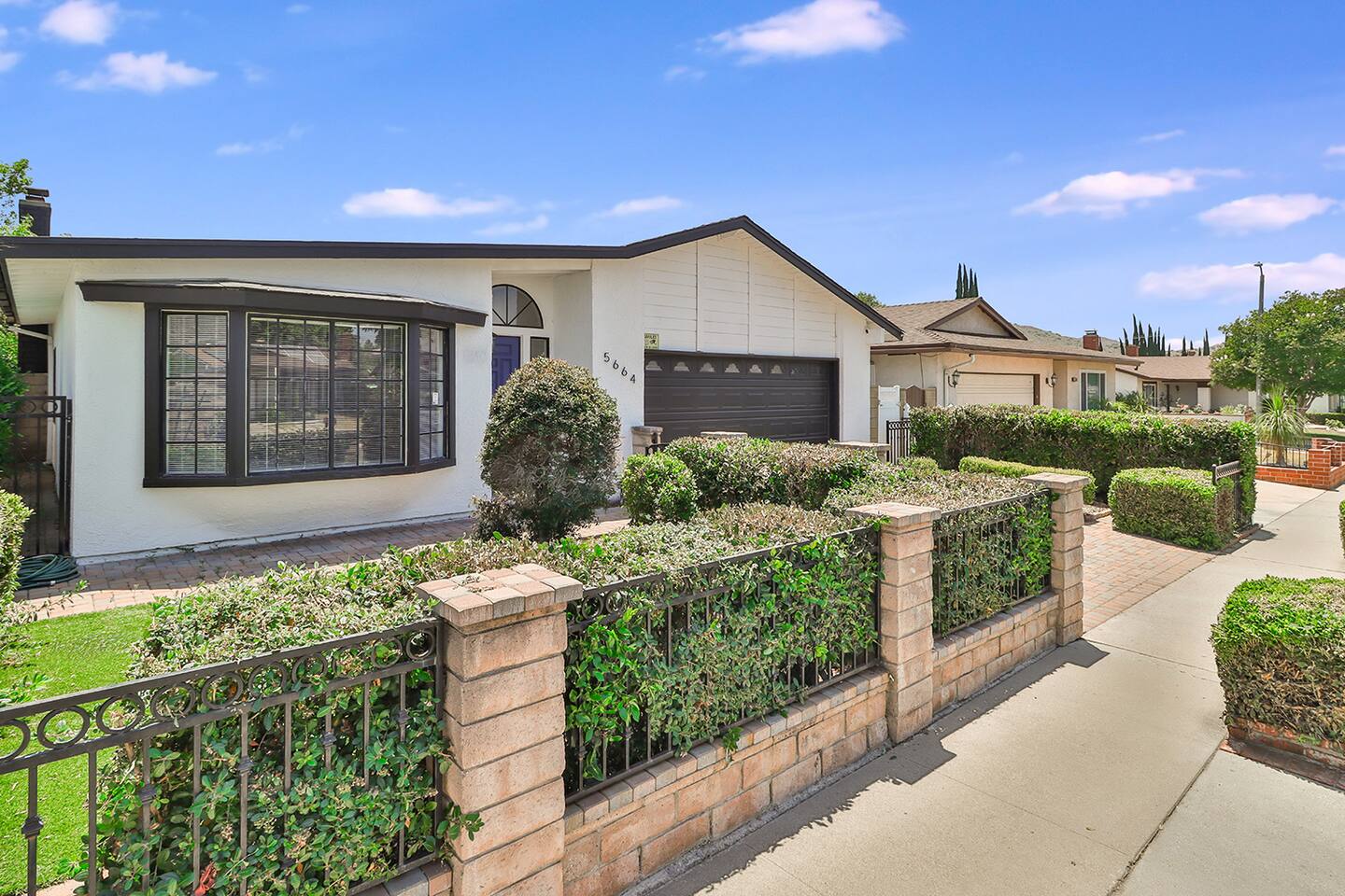 The exterior of a single-story home features a white facade with a dark roof, a prominent bay window, an attached garage, and a well-maintained front yard enclosed by a low brick and wrought-iron fence with lush green hedge.