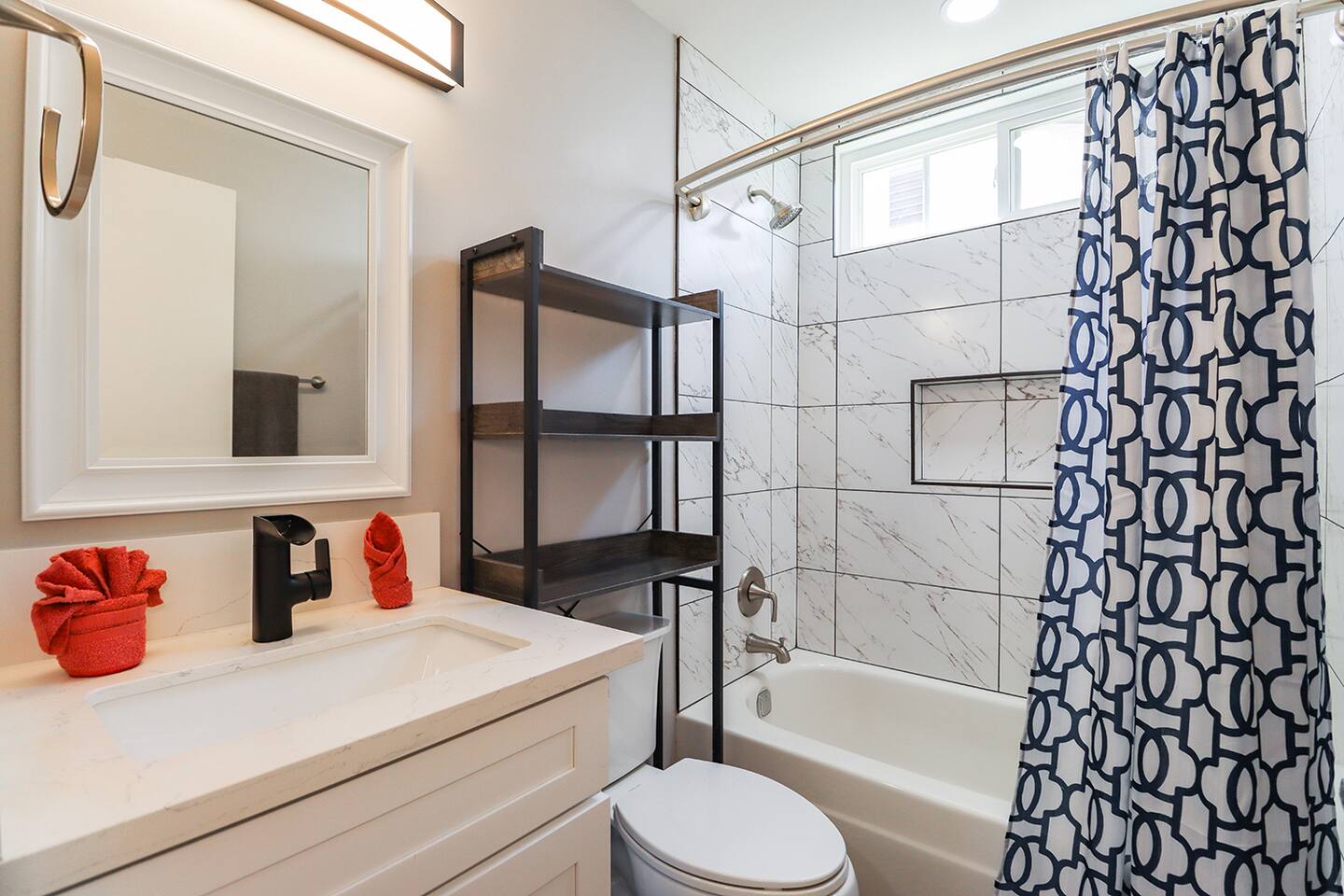 Bathroom presents a nice blend of modern and functional elements, featuring a sleek white vanity with a striking black faucet and a well-lit mirror, complemented by a conveniently placed dark metal shelving unit next to the toilet, and a bathtub area