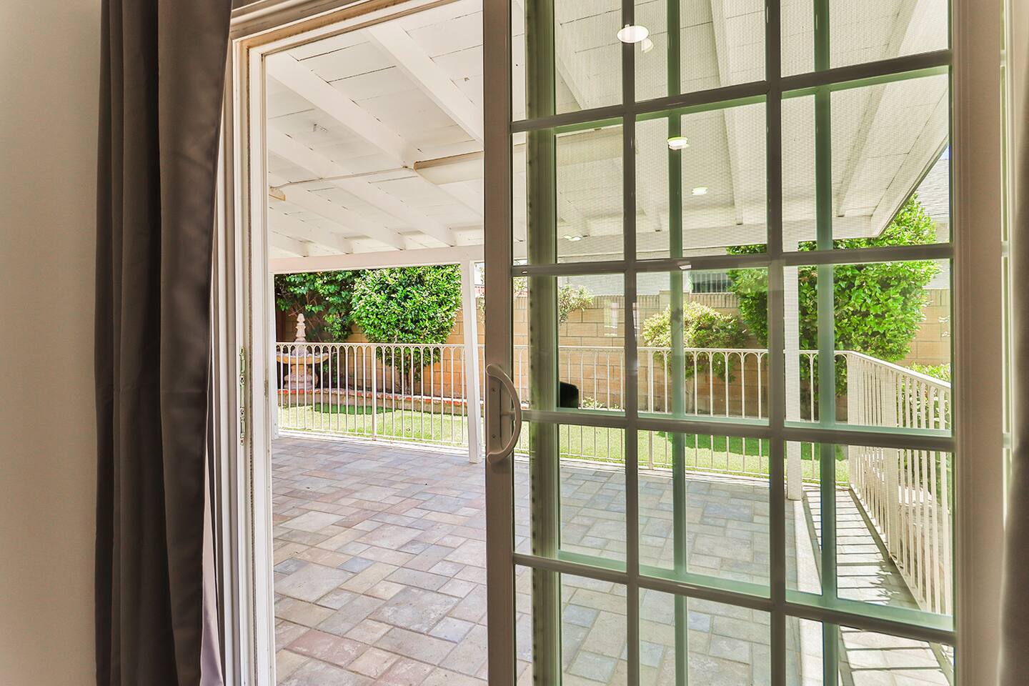 A view through partially opened sliding glass doors reveals a brick-paved patio area with white railings and lush green landscaping beyond, under a bright outdoor light.