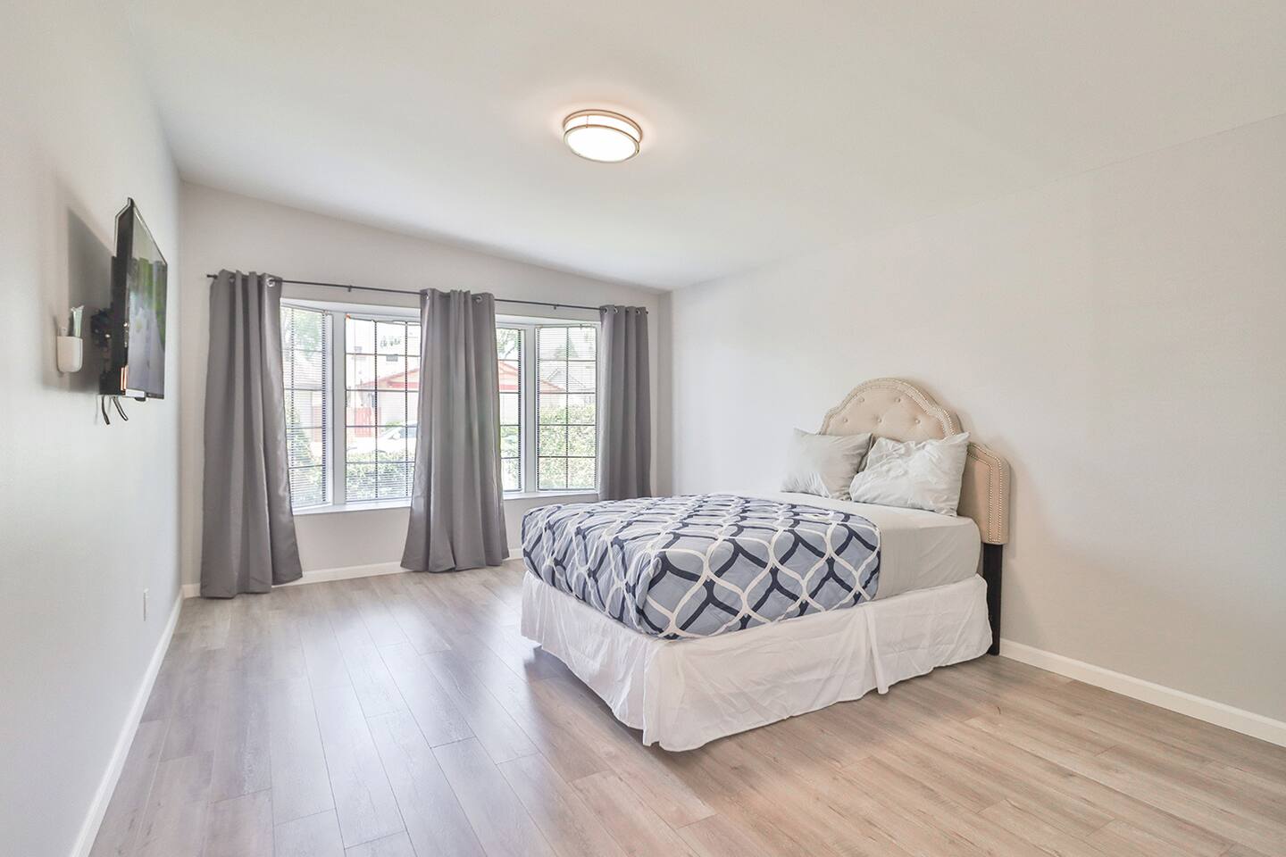 A bright bedroom features a bed with a gray and white patterned comforter and a beige headboard, positioned under a ceiling light and near two windows draped with gray curtains, with a mounted television on the adjacent wall.