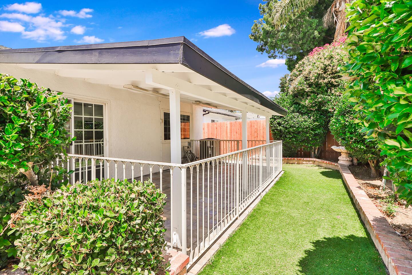 The exterior of a home showcases a covered patio with white railings, adjacent to a strip of artificial grass bordered by lush greenery.