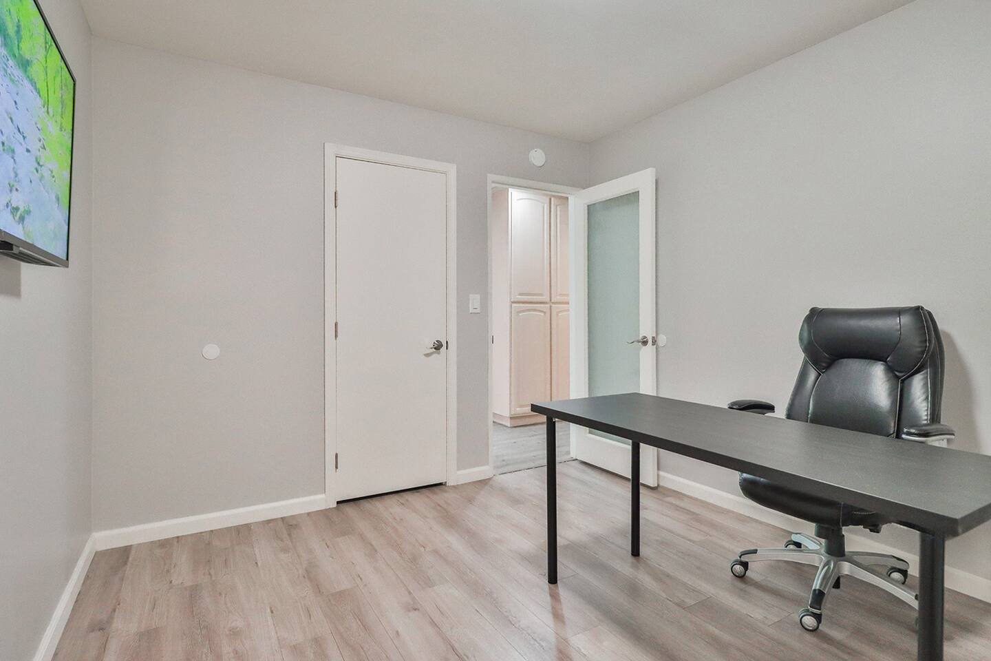 A minimalist home office writing table, a black ergonomic chair, a mounted television, and two white doors, set against light gray walls and wood-look flooring.