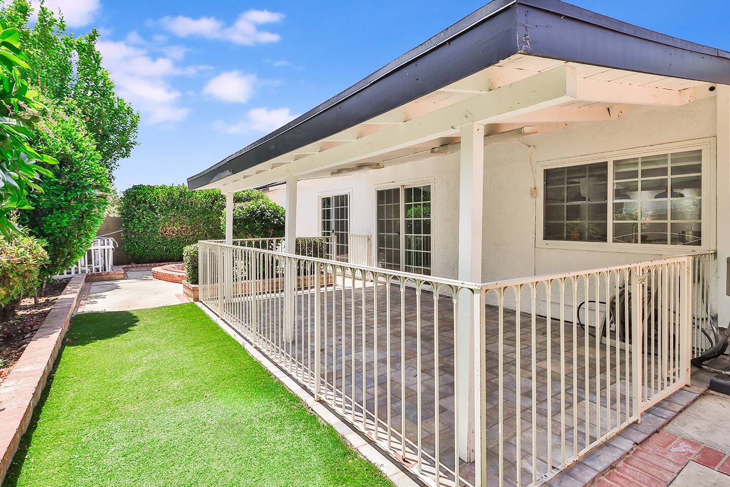 The exterior of a home features a covered patio with white railings and a brick-paved surface, adjacent to a strip of artificial turf and lush green shrubs.