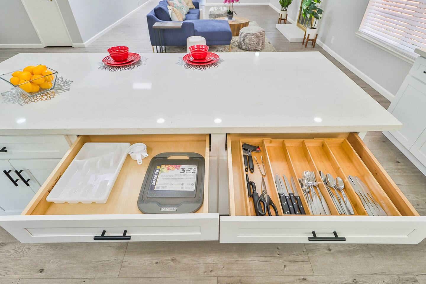 A kitchen island with a white countertop features two open drawers, one containing kitchen linens and a cutting board, and the other neatly organized with silverware and utensils.