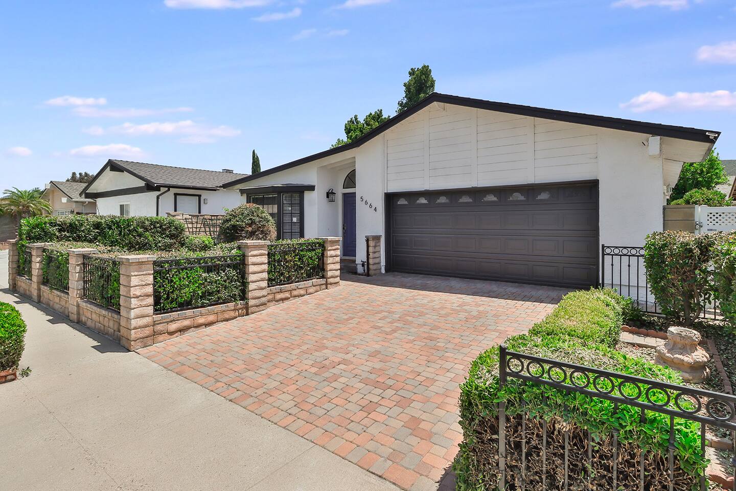 A single-story home's façade has a white front with a dark roof, a large bay window, an attached garage, and a well-kept front yard surrounded by a low brick and wrought-iron gate with a lush green hedge.