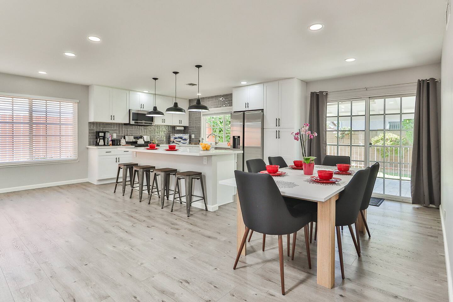 The open-concept dining area infeatures a light wood table with gray chairs and red place settings, positioned adjacent to a modern white kitchen with a central island and bar stools, all under recessed lighting and near sliding glass doors.