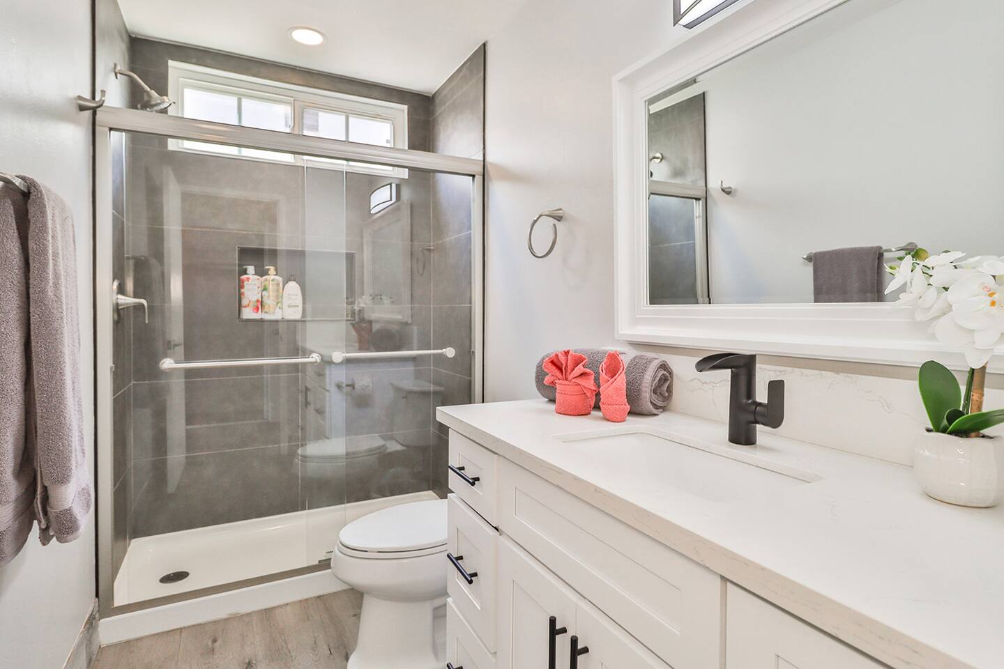 A modern bathroom features a glass-enclosed shower with gray tiled walls and a small window, a white vanity with a black faucet and a large mirror above, and a toilet.