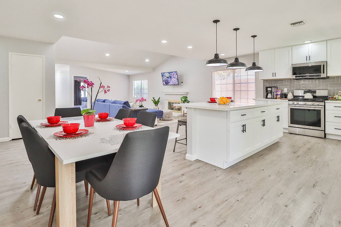The well-lit open-plan living space in  seamlessly integrates a dining area with a light wood table  a modern white kitchen featuring a central island and black pendant lights, and a comfortable living room.