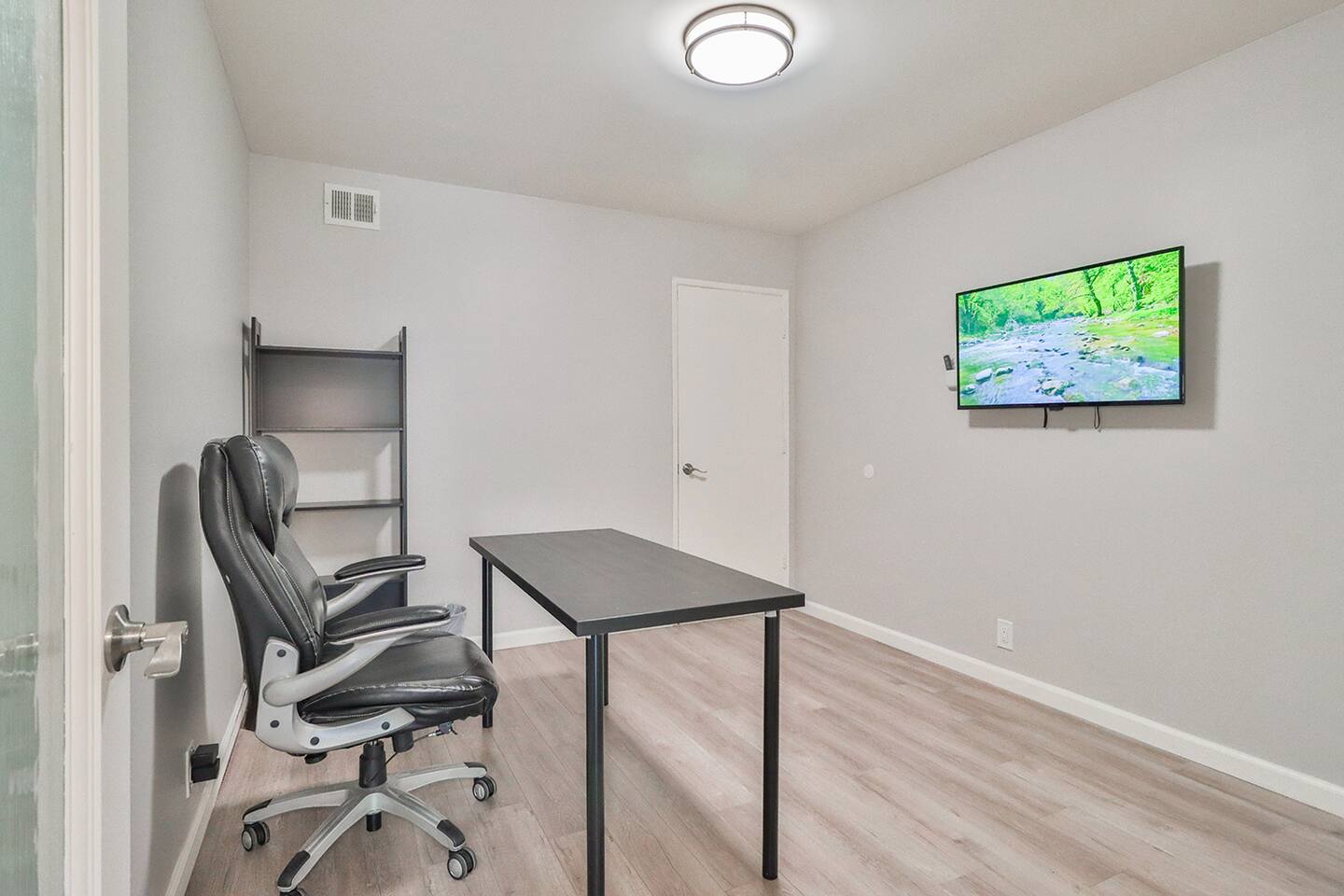 home office setup in a black desk, a black and gray ergonomic chair, a black shelving unit, and a mounted television displaying a nature scene, all within a room with light gray walls and wood-look flooring.