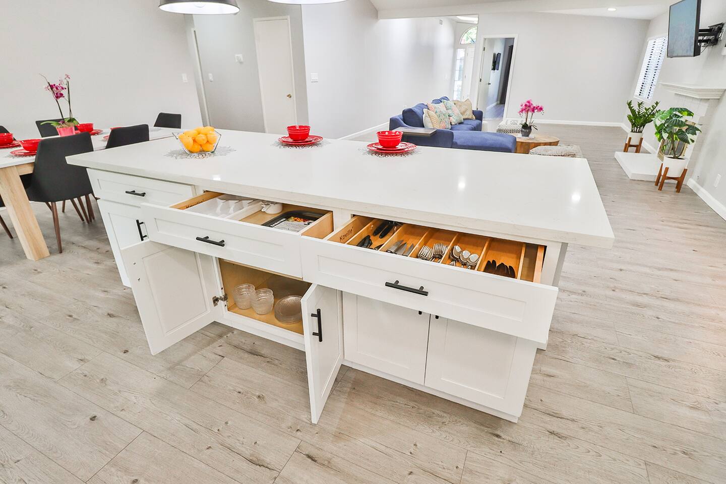 The bright kitchen island with its white countertop and cabinets, features open drawers revealing organized cutlery and utensils, all within an open-concept space that includes a dining area and living room.