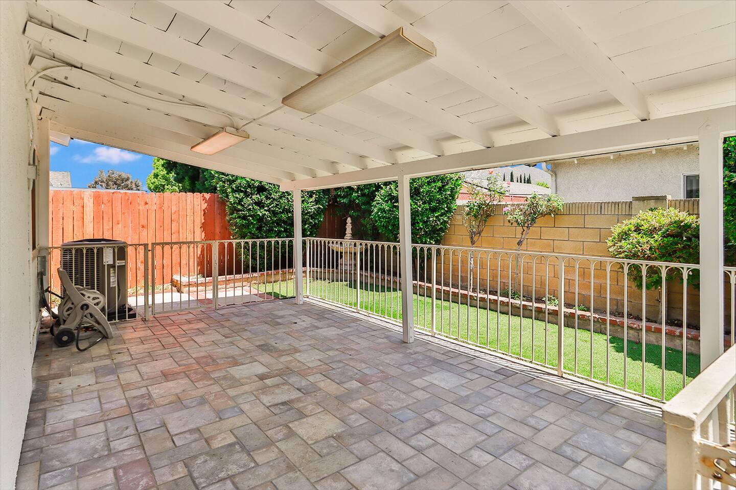 A covered patio features a brick paver floor, white support beams and ceiling, metal railings, and offers a view of a green lawn bordered by fences and lush greenery.