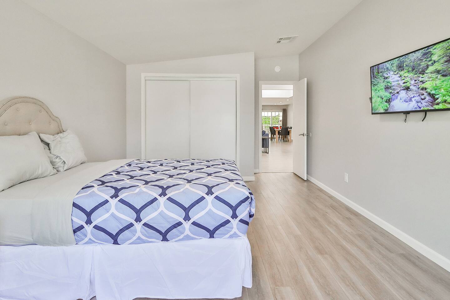 A simply furnished bedroom in contains a bed and a mounted television on the wall opposite an open doorway leading to another room.