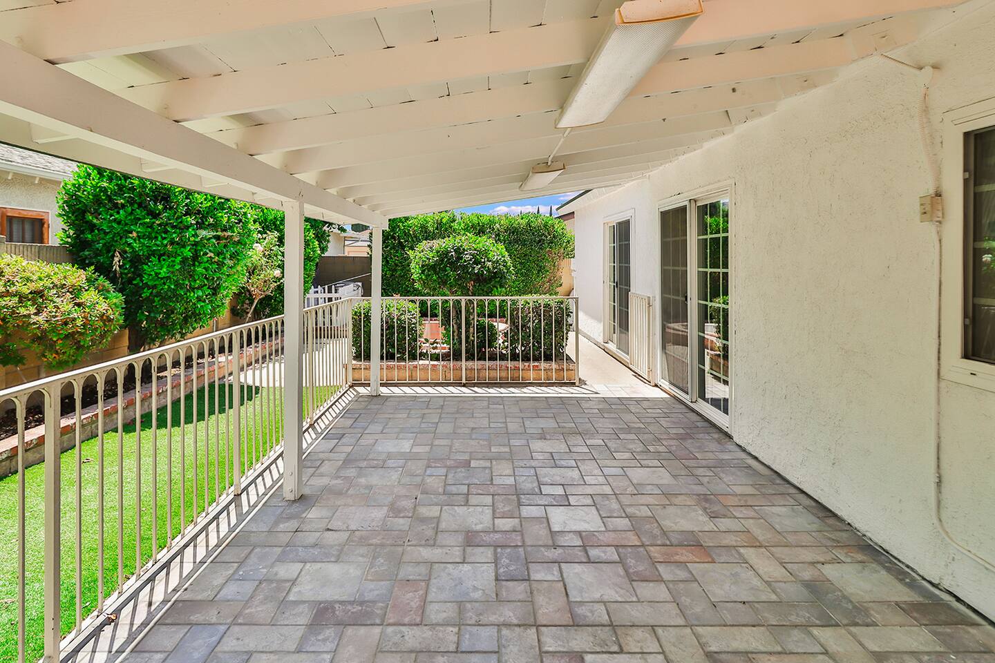 A covered patio features a brick paver floor, white support beams and ceiling, metal railings, and offers a view of a green lawn bordered by fences and lush greenery.
