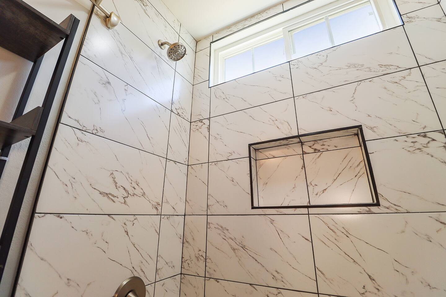 A detail of a bathtub and shower area in a bathroom showcases white tiles with gray veining, a showerhead, a small window above the tub, and a built-in niche for storage.