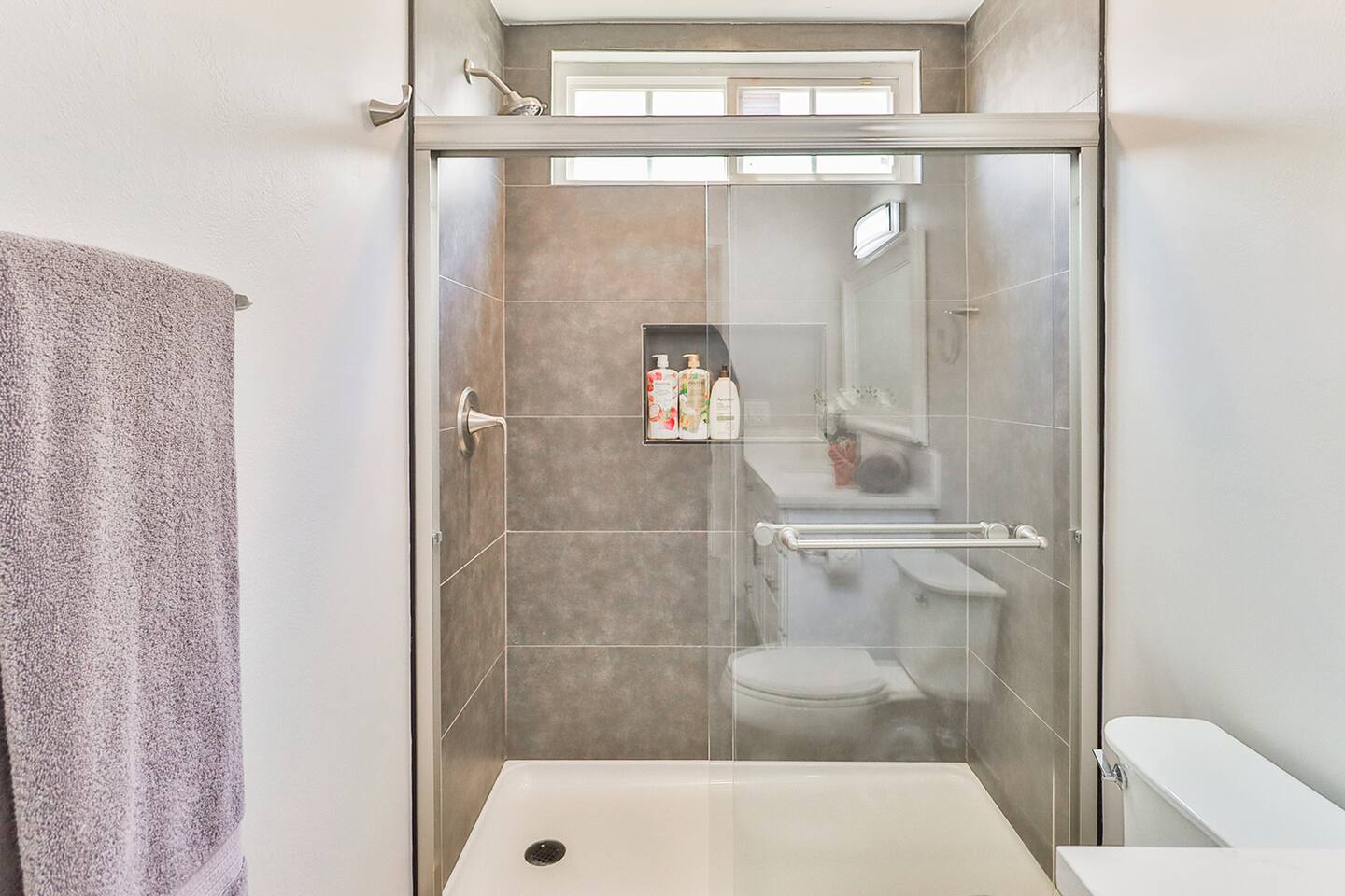 A modern shower in a bathroom features gray tiled walls with a built-in niche for toiletries, a glass sliding door, a showerhead, and a grab bar, with a partial view of a toilet visible through the glass.