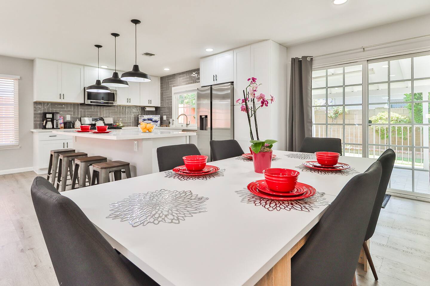 The dining area in features a white rectangular table set with red plates and bowls, surrounded by gray chairs, and situated near sliding glass doors that offer a view to the outside and connect it to the adjacent kitchen with a central island.