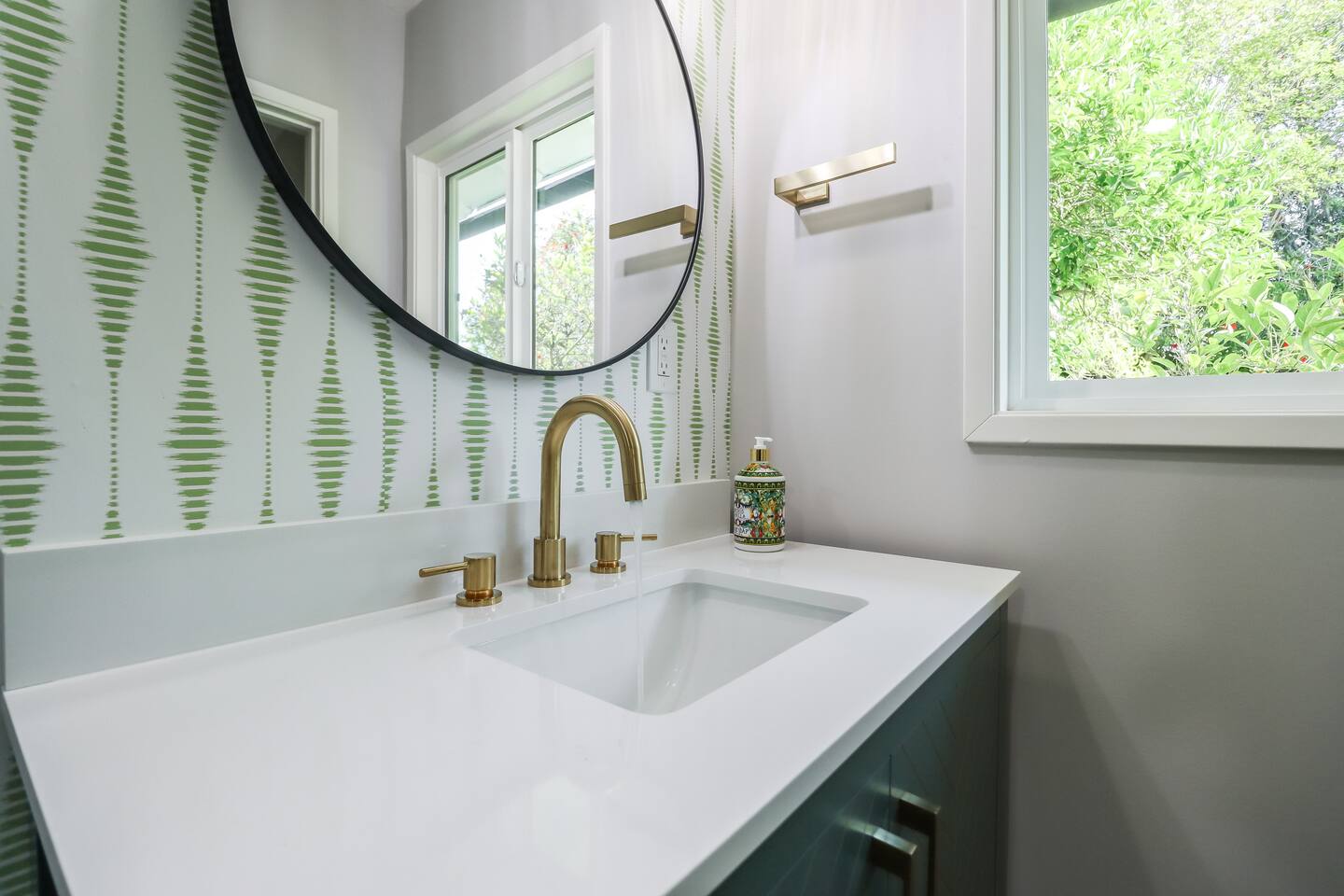 This modern bathroom features a stylish sage green vanity with gold chevron accents, a circular mirror, and a walk-in shower showcasing a mix of marble-style and green vertical subway tiles.