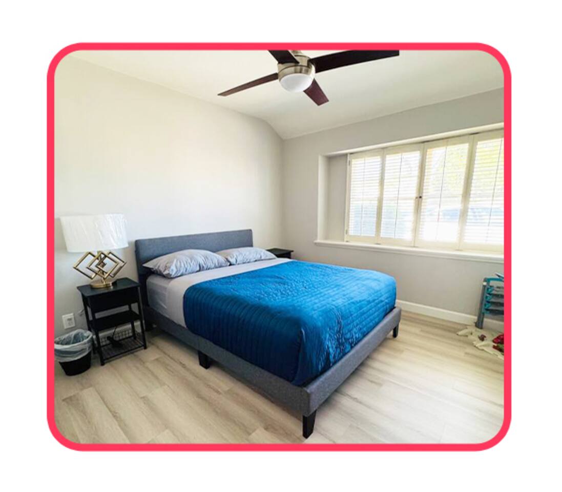 Comfortable bedroom featuring a queen-sized bed with a vibrant blue cover, designer gold lamp, and large window with plantation shutters.