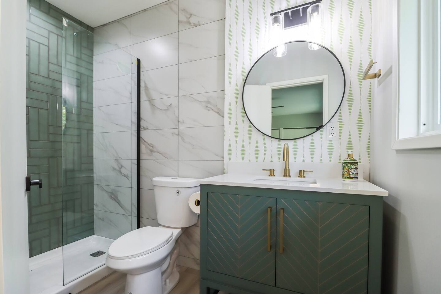 This modern bathroom features a stylish sage green vanity with gold chevron accents, a circular mirror, and a walk-in shower showcasing a mix of marble-style and green vertical subway tiles.