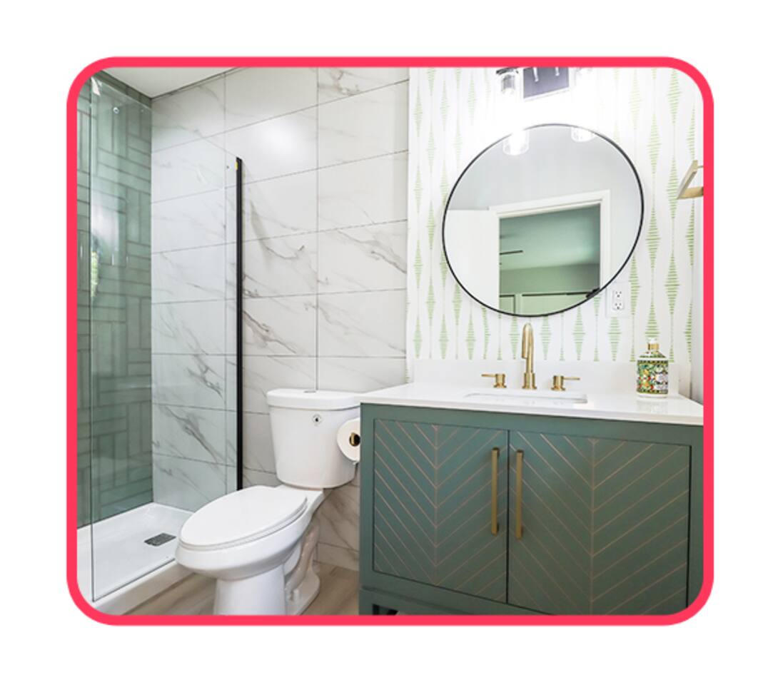 Designer bathroom featuring a sage green vanity with gold chevron detailing, white quartz countertop, brushed gold fixtures, a large circular mirror, and floor-to-ceiling marble-patterned shower tile.