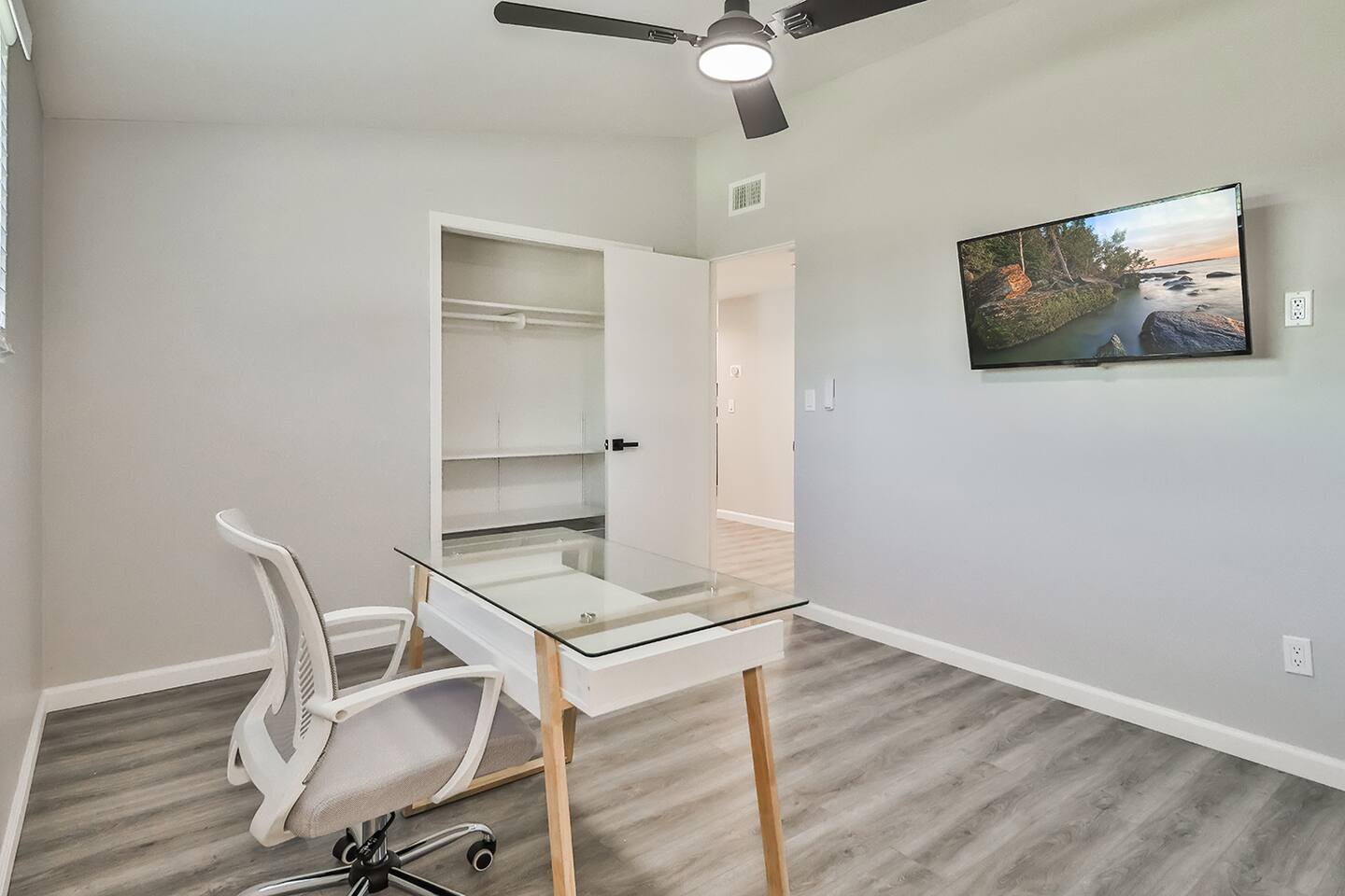 Functional workspace featuring a glass-top desk, ergonomic chair, wall-mounted TV, and an open closet with built-in shelving.