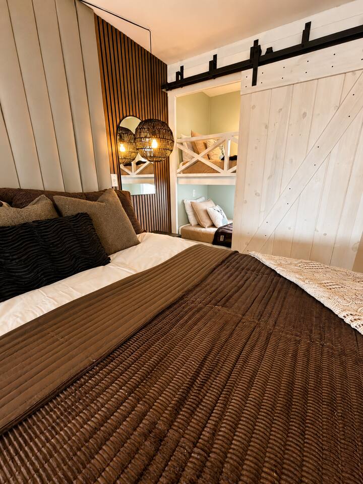 Relax in a beautifully designed bedroom featuring warm, cozy bedding, modern rustic accents, stylish lighting, and charming built-in bunks nearby—perfect for keeping the kids comfortably close while you unwind.