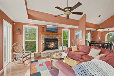 Living room with stone propane fireplace and TV above the mantel, twig rocking chair, pink sectional with yellow pillows and a gray knit throw, round coffee table, colorful geometric area rug, and an open view to the dining area.