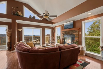 Living room with vaulted ceiling and clerestory windows, burgundy sectional sofa, round wooden coffee table, stone propane fireplace with a mounted TV above, ceiling fan, and sliding glass doors to a deck with mountain views.