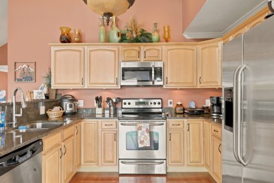 Kitchen with light wood cabinets, dark granite countertops, stainless steel range and oven, over-range microwave, side-by-side stainless refrigerator, dishwasher, double sink with a tall faucet, and decorative pottery above the cabinets.