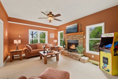 Den with beige carpet, brown leather sofa and armchair, wooden coffee table, ceiling fan, stone propane fireplace with a lit fire and TV above, and a yellow Pac-Man arcade cabinet standing beside a window.