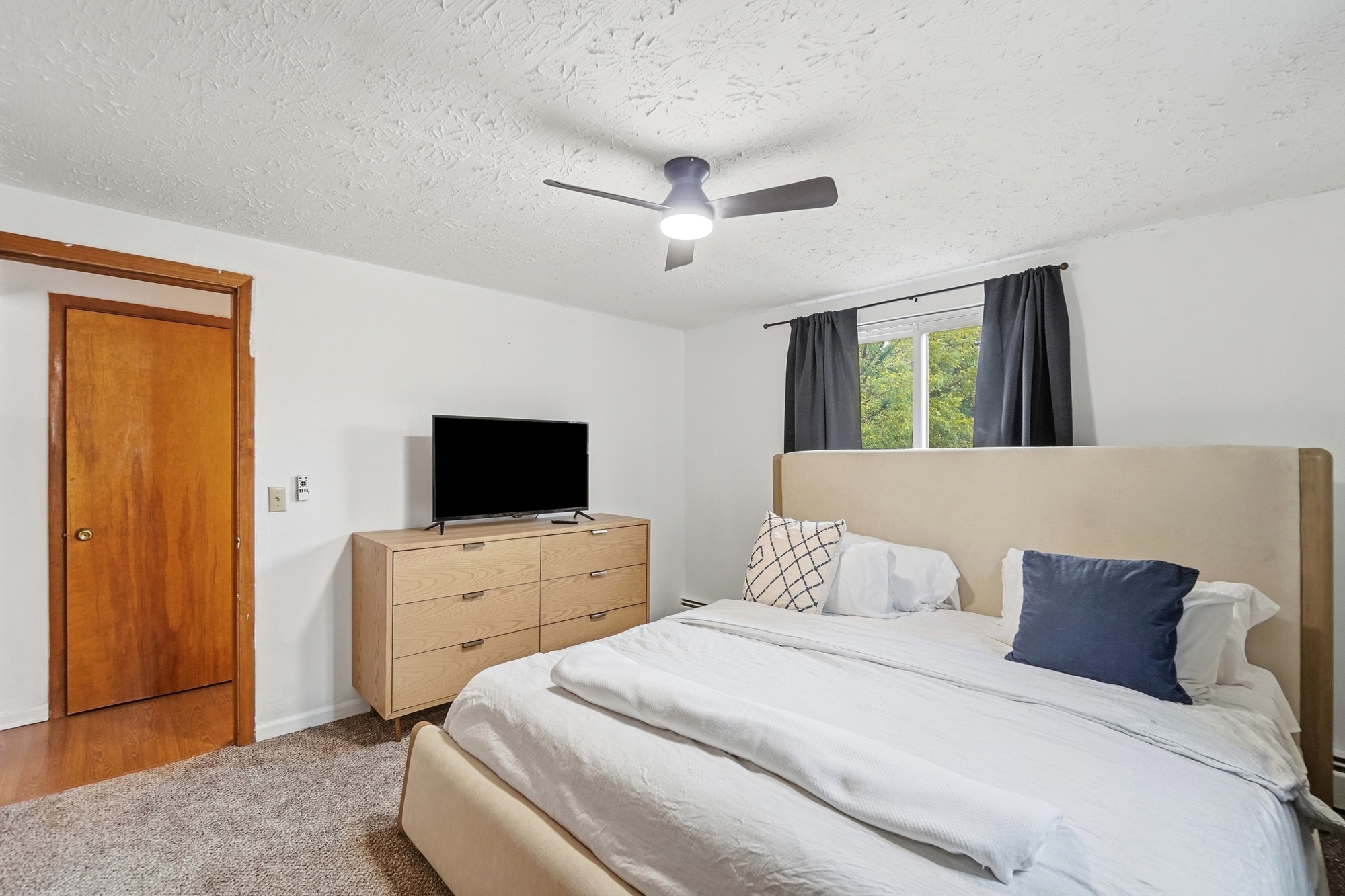 Bright, cozy bedroom with TV, ceiling fan, and plush upholstered bed